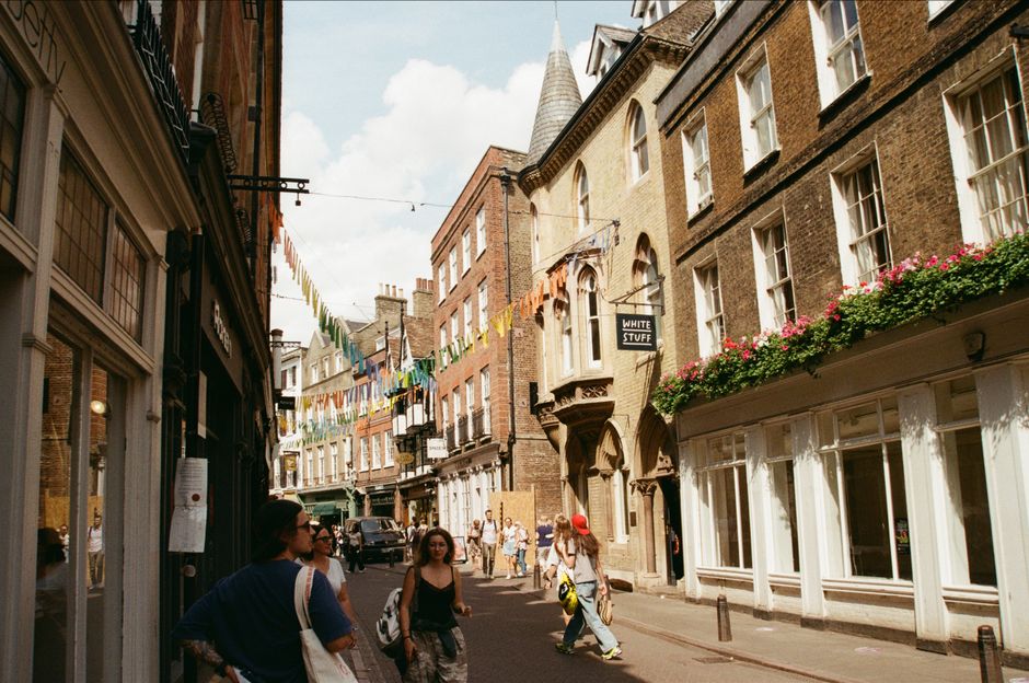 A narrow street scene in Spitalfields, showing historic brick and stone buildings with large windows decorated with flower boxes filled with pink and green foliage. Colourful triangular bunting flags