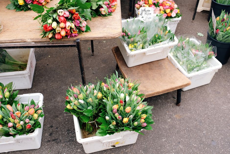 At a flower market in Spitalfields, several white plastic crates and wooden tables display freshly cut tulip bouquets in various stages of arrangement. The tulips are in shades of pink, orange, white,