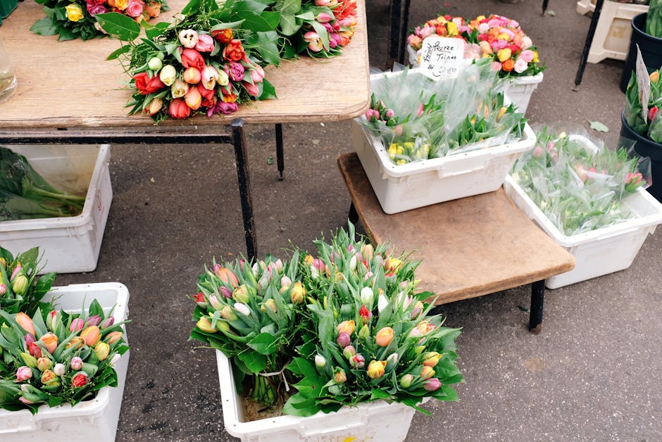 A collection of fresh tulip bouquets at an outdoor flower market shelf, featuring pink, yellow, white, and purple blooms with lush green leaves, arranged in white plastic containers and wrapped in cle