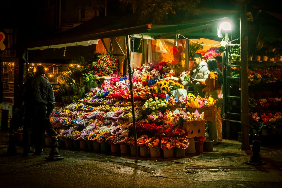 Photograph of a vibrant floral market stall at night, illuminated by warm artificial lighting, showcasing an abundant display of fresh flowers including sunflowers, roses, lilies, and carnations in va