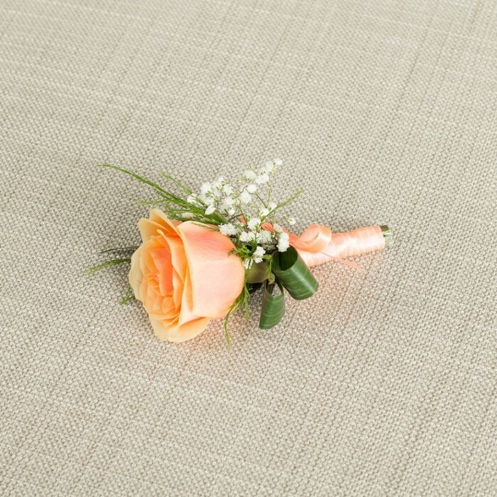 A single peach rose boutonniere is captured resting horizontally on a light, oatmeal-beige fabric with a clear linen-style weave, each thread forming a soft grid that offers understated texture without competing with the flowers. The rose is partially open, its petals forming a spiralled heart that deepens into a warm, glowing peach with hints of apricot, then lightening gradually towards the outer edges into a creamy, almost ivory blush. It has the fresh, velvety look of a bloom cut that morning from a well-tended garden not far from Spitalfields' cobbled streets. Clustered just above and beside the rose, tiny white baby's breath blossoms add airiness and contrast, their minuscule petals catching the light like scattered pearls. Slightly behind, slender green blades and a couple of polished, broader leaves emerge in varying tones of green, giving the arrangement shape and a gentle sense of movement, as though a breeze from nearby Allen Gardens had brushed through. The stems are carefully aligned and wrapped with a smooth peach satin ribbon that closely matches the rose, the ribbon laid in snug, even coils and finished with a modest bow or tucked knot that keeps the design streamlined for a suit lapel. Soft, even lighting falls across the scene, highlighting the natural sheen of the petals, the cloud-like gypsophila, and the fine texture of the fabric ground, creating an image that feels calm and intimate. This boutonniere suggests a moment just before a ceremony in Spitalfields-perhaps outside a converted warehouse venue-where a groom, partner, or cherished guest is about to pin on a small emblem of joy, romance, and thoughtful craftsmanship.
