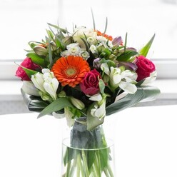 Inspired by the creative spirit and heritage of Spitalfields, this exquisite bouquet unites bold colours, varied textures, and modern artistry. At its centre, radiant gerbera daisies-two fiery orange, two soft peach-fan out like lanterns, their velvety petals encircling dark, inviting centres that draw the gaze. Intertwined with these are plush, fuchsia-tinged roses, their tightly arranged petals adding drama and opulence, balanced by sprays of pristine white alstroemeria and petite white filler blossoms reminiscent of local markets' floral stalls. Underneath, a flourish of glossy, emerald-green leaves provides a grounding contrast to the petals' brilliance, broad aspidistra leaves wrapping the stems much as Spitalfields' famed historic walls embrace the area's creativity. Visual interest is heightened by subtle pink blooms and various slender greens that add volume and a lively interplay of light and shadow. The clear glass vase, with its visible, verdant stems, speaks to freshness and authenticity-qualities long associated with Spitalfields. Bathed in luminous, diffused daylight, the arrangement sings with a cheerful energy-perfect for celebratory occasions, milestone events, or simply to uplift a loved one's home in the lively lanes of Spitalfields.